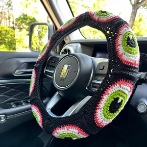 May include: A black, white, and green crocheted steering wheel cover with a repeating eye pattern.