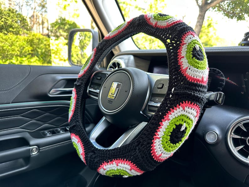 May include: A black, white, and green crocheted steering wheel cover with a repeating eye pattern.