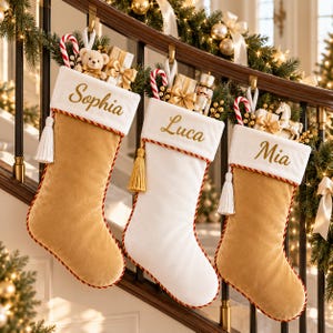 Puede incluir: Tres calcetines de Navidad con puños blancos bordados con los nombres Sophia, Luca y Mia. Los calcetines son de terciopelo dorado y blanco, adornados con cordón trenzado rojo y dorado y borlas blancas, rellenos de bastones de caramelo y juguetes.