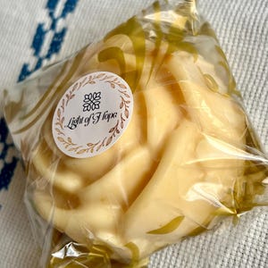 Può includere: Una saponetta a forma di rosa, di colore giallo chiaro, è confezionata in cellophane trasparente con dettagli dorati. Un adesivo circolare con la scritta "Light of Hope" è sul davanti. Il sapone è di colore giallo pallido.