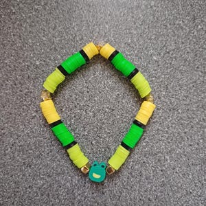 Peut inclure: Un bracelet en perles vertes et jaunes avec un charme de grenouille. Le bracelet est composé de perles rondes colorées avec des rayures noires.