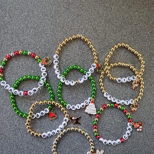 May include: A collection of eight Christmas-themed beaded bracelets with different words and charms. The bracelets are made with green, red, and gold beads and feature charms such as a Christmas tree, a candy cane, a bell, and a snowflake.