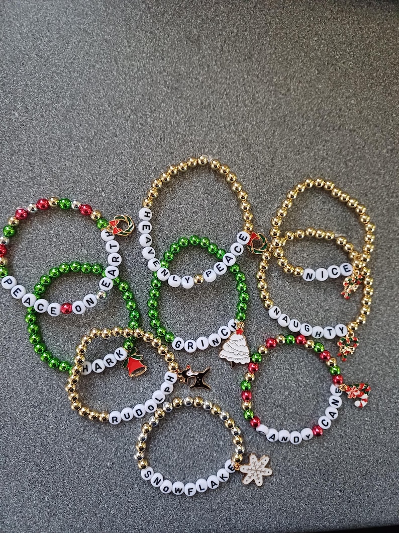 May include: A collection of eight Christmas-themed beaded bracelets with different words and charms. The bracelets are made with green, red, and gold beads and feature charms such as a Christmas tree, a candy cane, a bell, and a snowflake.