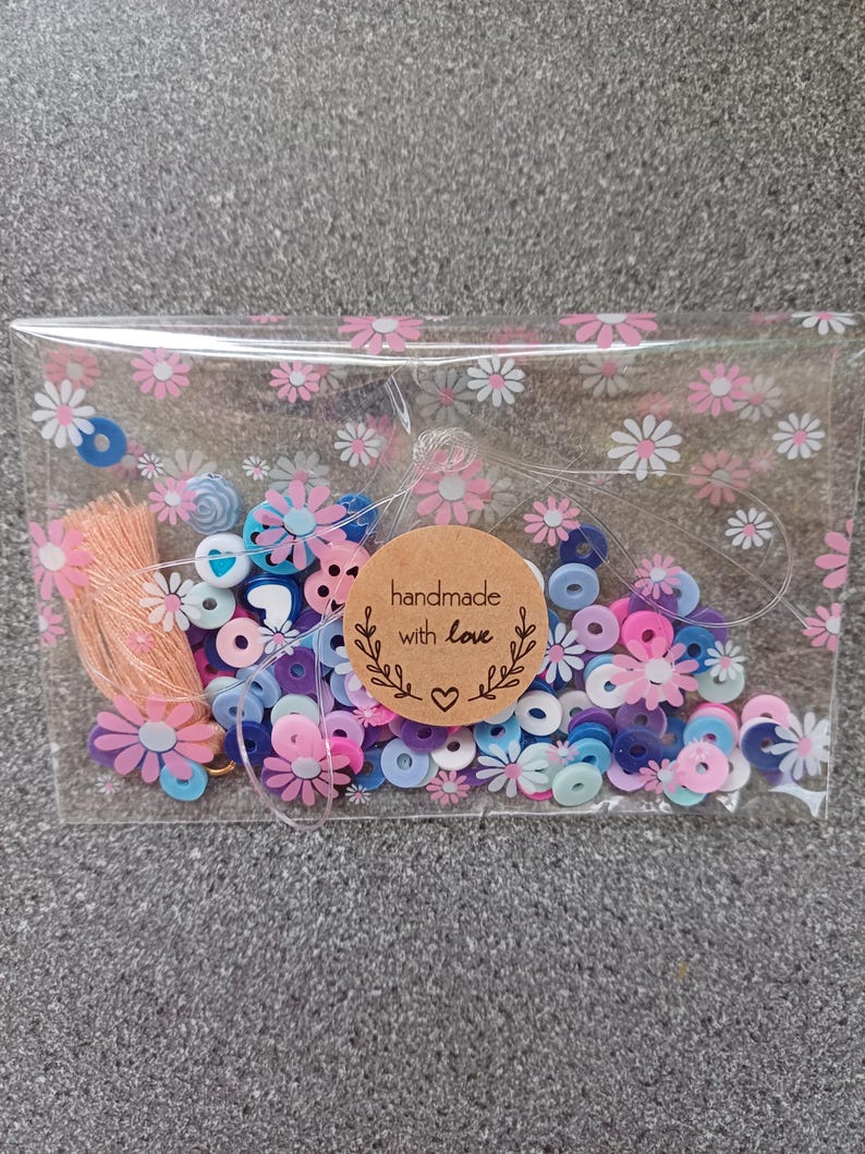 May include: A clear plastic bag filled with colorful beads and small flowers. The bag has a pink and white floral pattern. A brown paper tag with the text "handmade with love" is attached to the bag.
