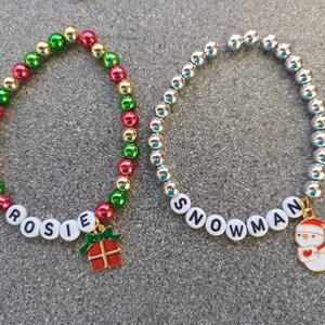 May include: Two beaded bracelets with charms. One bracelet is red, green, and gold with the word "ROSIE" and a gift box charm. The other bracelet is silver with the word "SNOWMAN" and a snowman charm.