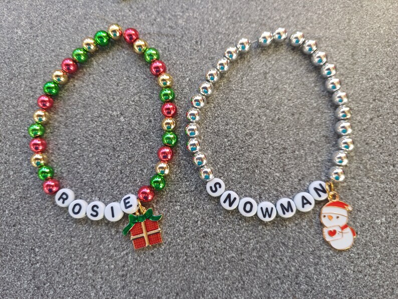 May include: Two beaded bracelets with charms. One bracelet is red, green, and gold with the word "ROSIE" and a gift box charm. The other bracelet is silver with the word "SNOWMAN" and a snowman charm.