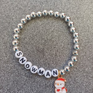 May include: A silver beaded bracelet with white letter beads spelling "SNOWMAN" and a gold charm of a snowman with a red heart.