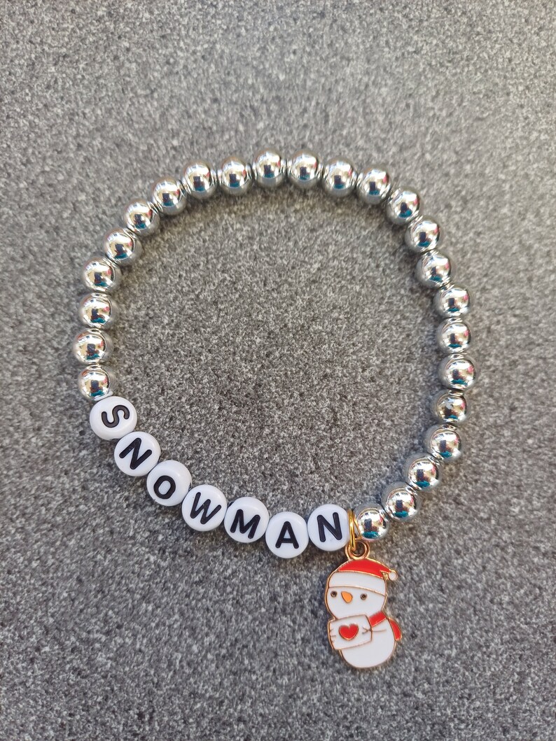 May include: A silver beaded bracelet with white letter beads spelling "SNOWMAN" and a gold charm of a snowman with a red heart.