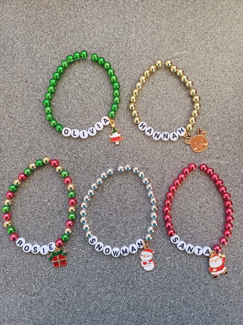 May include: Five Christmas-themed beaded bracelets with name charms and festive charms. The bracelets are green, gold, red, silver, and red and green. The charms include a Christmas tree, reindeer, snowman, Santa Claus, and a gift box.
