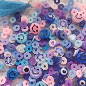 May include: A variety of colorful plastic beads, including smiley faces, rings, and evil eyes. The beads are in shades of blue, pink, purple, and white.