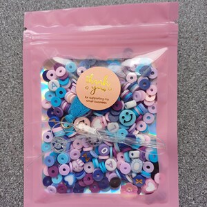 May include: A pink resealable bag filled with colorful beads and charms. The beads are in shades of blue, purple, and pink. Some of the charms are shaped like hearts, smiley faces, and letters. A sticker on the bag says "Thank you! for supporting my small business."