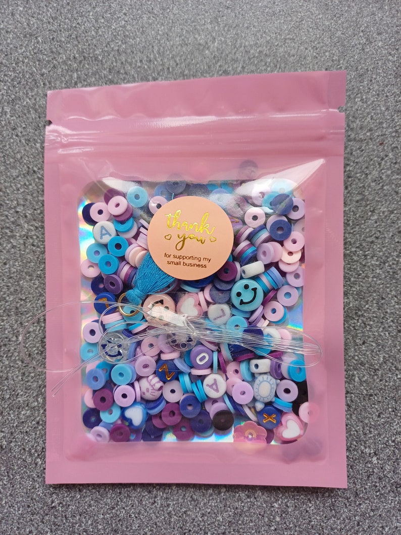 May include: A pink resealable bag filled with colorful beads and charms. The beads are in shades of blue, purple, and pink. Some of the charms are shaped like hearts, smiley faces, and letters. A sticker on the bag says "Thank you! for supporting my small business."