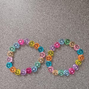 May include: Two beaded bracelets with smiley face beads in a variety of colors, including pink, orange, yellow, green, blue, and purple. The bracelets are arranged side by side on a gray surface.