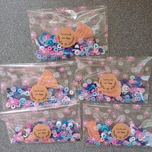 May include: Five clear plastic bags with pink floral patterns contain colorful paper beads, including blue, pink, white, and purple. Each bag has a brown paper tag with the text "handmade with love" and a small pink heart.