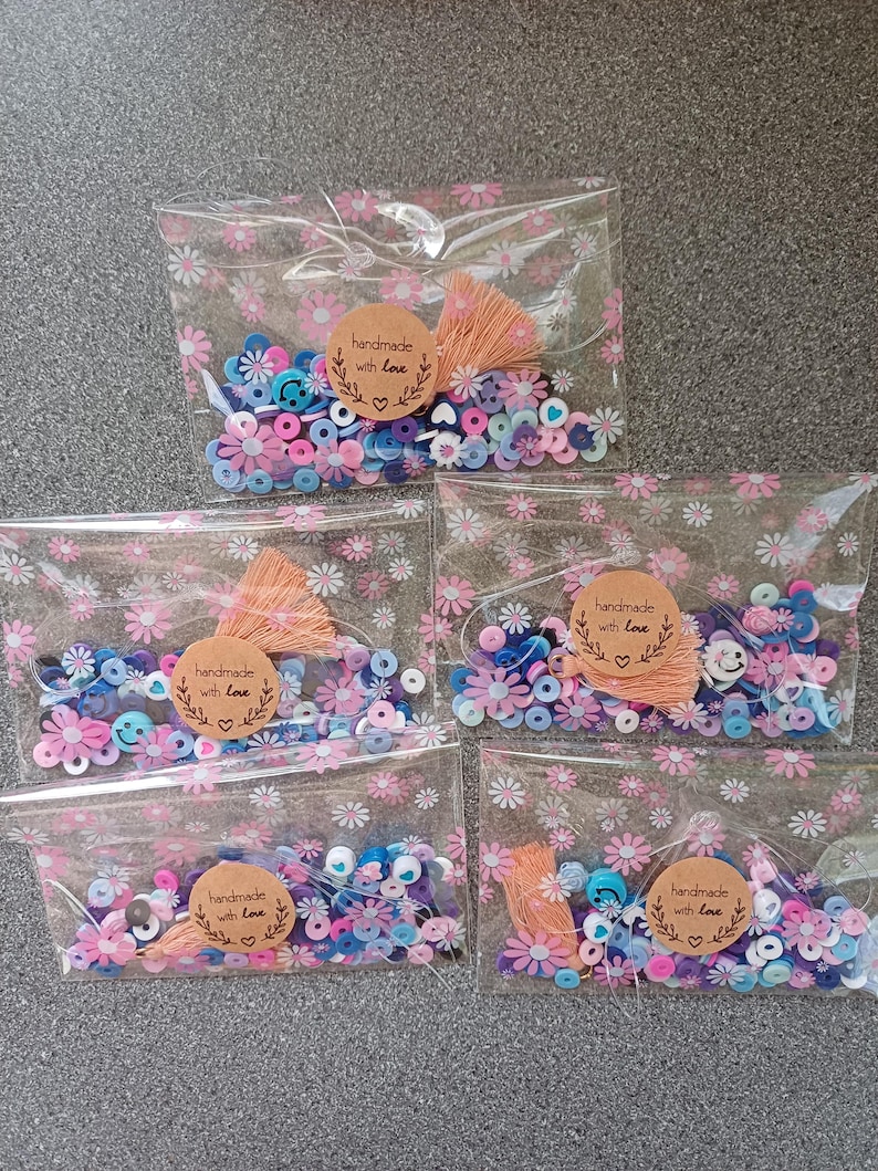 May include: Five clear plastic bags with pink floral patterns contain colorful paper beads, including blue, pink, white, and purple. Each bag has a brown paper tag with the text "handmade with love" and a small pink heart.