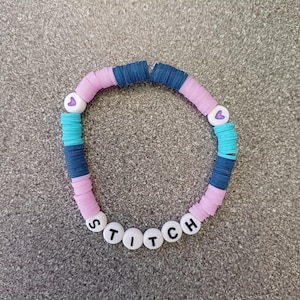 Könnte beinhalten: Ein farbenfrohes Perlenarmband mit rosa, blauen und weißen Perlen. Die Perlen buchstabieren das Wort "STITCH" mit weißen Buchstaben auf schwarzen Perlen.