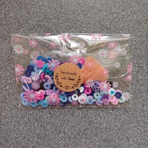 May include: A clear plastic bag filled with colorful beads and a small brown tag that says "handmade with love". The beads are in shades of pink, blue, and purple, and some have a flower or heart shape.