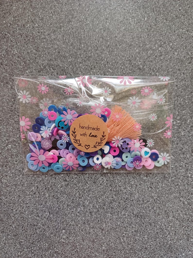 May include: A clear plastic bag filled with colorful beads and a small brown tag that says "handmade with love". The beads are in shades of pink, blue, and purple, and some have a flower or heart shape.