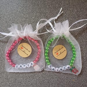 May include: Two Christmas-themed bracelets in organza bags. The red bracelet has white beads that spell "SANTA" and a small Santa Claus charm. The green bracelet has white beads that spell "GRINCH" and a small Grinch charm. Both bracelets have a small round tag that says "handmade with love".