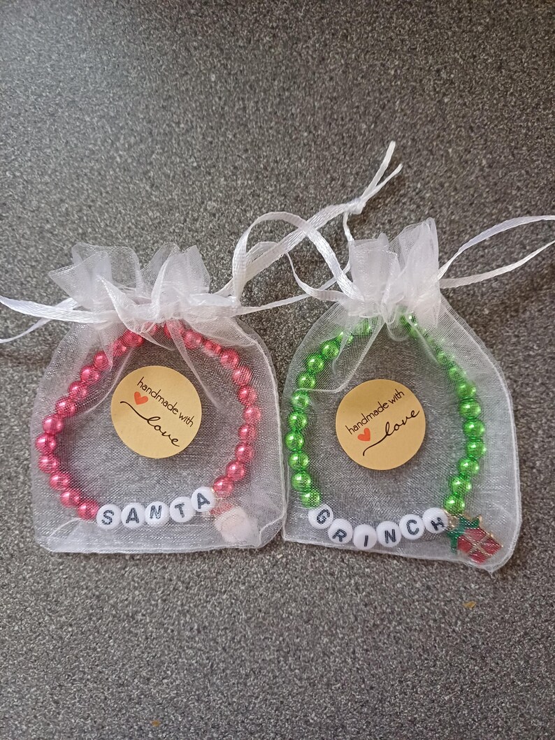 May include: Two Christmas-themed bracelets in organza bags. The red bracelet has white beads that spell "SANTA" and a small Santa Claus charm. The green bracelet has white beads that spell "GRINCH" and a small Grinch charm. Both bracelets have a small round tag that says "handmade with love".