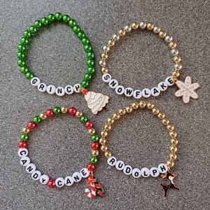 May include: Four Christmas themed beaded bracelets with charms. The bracelets are green, gold, red and gold, and gold. The charms are a Christmas tree, a snowflake, a candy cane and a reindeer.