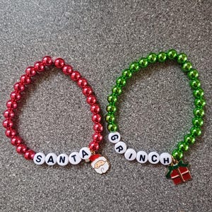 May include: Two beaded bracelets with the words "SANTA" and "GRINCH" spelled out in white beads. The "SANTA" bracelet is red and has a Santa Claus charm. The "GRINCH" bracelet is green and has a Grinch charm with a gift.