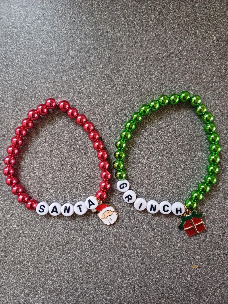 May include: Two beaded bracelets with the words "SANTA" and "GRINCH" spelled out in white beads. The "SANTA" bracelet is red and has a Santa Claus charm. The "GRINCH" bracelet is green and has a Grinch charm with a gift.