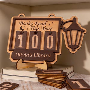 Könnte beinhalten: Ein Bücherzähler aus Holz mit dem Text "Books Read This Year" und "Olivia's Library". Der Zähler zeigt die Zahl "100" an. Zusätzliche Holznummernkacheln sind enthalten. Eine dekorative Laterne ist an der Seite befestigt.