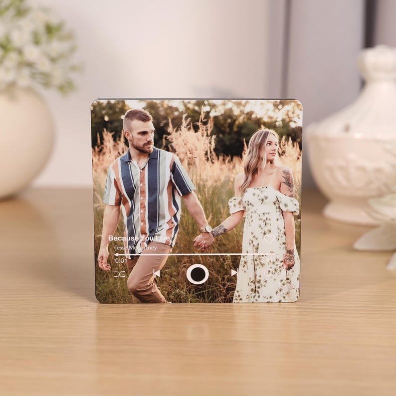 Pode incluir: Um bloco de madeira quadrado com uma fotografia de um casal de m&atilde;os dadas. A imagem &eacute; projetada para se assemelhar a uma interface de reprodutor de m&uacute;sica, com o texto "Because You Love" e informa&ccedil;&otilde;es do artista. O bloco est&aacute; sobre uma superf&iacute;cie de madeira.