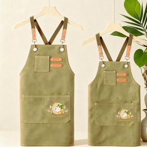 May include: Two olive green aprons with brown leather details and pockets. The aprons feature adjustable straps and a floral design with the text "Alice's kitchen". They are hanging on wooden hangers.