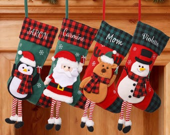 May include: Four Christmas stockings hang from a wooden mantel. Each stocking is green or red with a plaid cuff and features a different character: a penguin, Santa, a reindeer, and a snowman. Names are embroidered on the cuffs.
