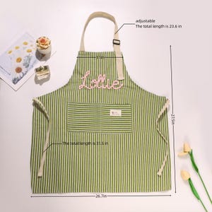 K&ouml;nnte beinhalten: Gr&uuml;n-wei&szlig; gestreifte Sch&uuml;rze mit dem Namen "Lollie" in Rosa gestickt. Die Sch&uuml;rze hat einen verstellbaren Riemen und eine Tasche. Die Gesamtl&auml;nge betr&auml;gt 80 cm, die verstellbare L&auml;nge 60 cm.