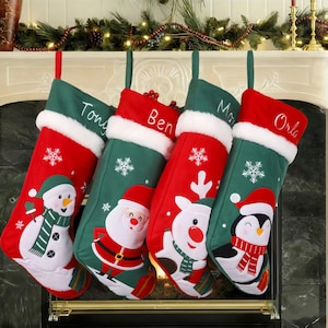 Könnte beinhalten: Vier Weihnachtsstrümpfe mit roten und grünen Designs hängen an einem Kamin. Jeder Strumpf hat eine weiße Manschette und zeigt einen anderen Weihnachtscharakter: einen Schneemann, den Weihnachtsmann, ein Rentier und einen Pinguin. Namen sind auf den grünen Oberteilen gestickt.
