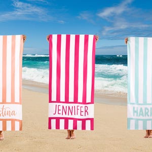 Könnte beinhalten: Drei Strandtücher mit vertikalen Streifen in Koralle, Pink und Hellblau, jedes mit einem weißen Band am unteren Rand. Die Handtücher sind mit den Namen "Christina", "Jennifer" und "Harper" in Schreibschrift personalisiert. Der Hintergrund ist eine Strandszene.