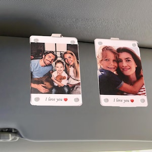 Peut inclure: Deux supports rectangulaires blancs pour visière de voiture avec des photos de famille. Chaque support a un clip métallique et le texte "I love you" avec un cœur. Une photo montre une famille de trois personnes, l'autre une mère et son enfant.
