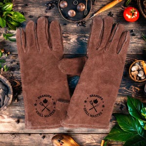 Könnte beinhalten: Ein Paar braune Leder-Grillhandschuhe. Auf jedem Handschuh steht in schwarzer Schrift "GRANDPA GRILL MASTER", zusammen mit einer Grafik einer Flamme, gekreuzten Zangen und einer Gabel. Die Handschuhe liegen auf einer Holzoberfläche.