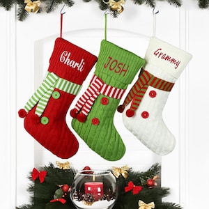 Könnte beinhalten: Drei personalisierte Weihnachtsstrümpfe aus rotem, grünem und weißem Strickstoff. Die Strümpfe haben einen weißen Pomponbesatz und einen rot-weiß gestreiften Schal. Die Strümpfe sind mit den Namen "Charli", "Josh" und "Grammy" personalisiert.
