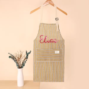 K&ouml;nnte beinhalten: Gestreifte Sch&uuml;rze in Braun und Wei&szlig; mit dem Namen "Elias" in rosa Schrift. Die Sch&uuml;rze hat eine gro&szlig;e Vordertasche und h&auml;ngt an einem Holzb&uuml;gel. Ein kleines Etikett ist auf der Tasche sichtbar.