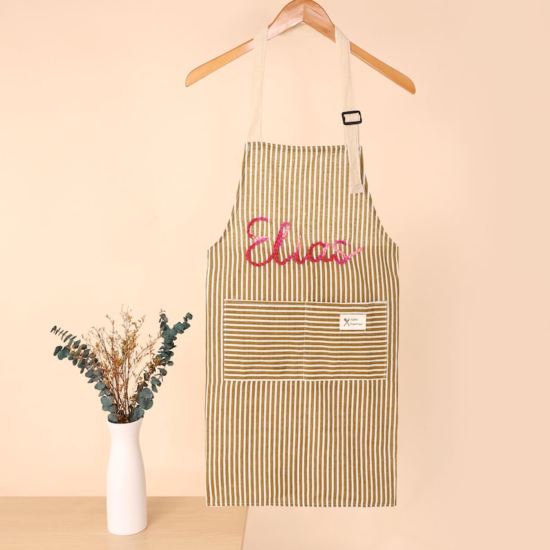 K&ouml;nnte beinhalten: Gestreifte Sch&uuml;rze in Braun und Wei&szlig; mit dem Namen "Elias" in rosa Schrift. Die Sch&uuml;rze hat eine gro&szlig;e Vordertasche und h&auml;ngt an einem Holzb&uuml;gel. Ein kleines Etikett ist auf der Tasche sichtbar.