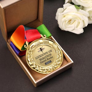 May include: A gold-coloured circular medal with a laurel wreath border, featuring the text LOCKDOWN 10K MARATHON, Daniel Roche, and 2025. It includes a multi-coloured ribbon in orange, red, green, and blue, presented inside a tan-lined display box.