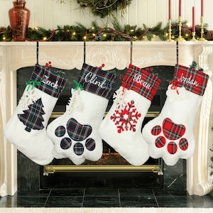 May include: Four personalized Christmas stockings with white fleece, red and green plaid trim, and paw print designs. The stockings are hung on a fireplace mantel and are labeled "Zack", "Clint", "Beau", and "Jordan".
