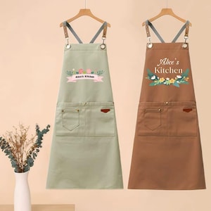Könnte beinhalten: Zwei Schürzen hängen an Holzbügeln. Eine ist salbeigrün mit einem rosa Banner mit der Aufschrift "Alice's Kitchen" und floralen Akzenten. Die andere ist braun mit demselben Text und floralen Akzenten. Beide haben Taschen und Lederakzente.