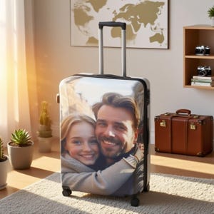 May include: A black rolling suitcase featuring a photograph of a smiling couple. The suitcase has a retractable handle and four wheels. A brown leather suitcase and two vintage cameras are visible in the background.