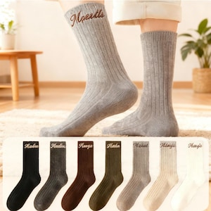 Peut inclure: Deux chaussettes en tricot côtelé gris, l'une avec le mot "Moentls" brodé en marron. En dessous, six paires de chaussettes en noir, gris foncé, marron, vert olive, gris clair, crème et blanc, chacune avec un nom brodé différent.