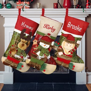 Könnte beinhalten: Drei rot-grüne Weihnachtsstrümpfe mit den bestickten Namen "Mommy", "Kirby" und "Brooke". Jeder Strumpf zeigt einen anderen festlichen Charakter: ein Rentier, einen Schneemann und den Weihnachtsmann.