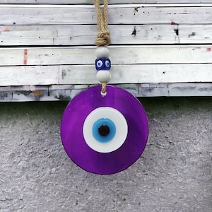 May include: Purple evil eye charm with a white and blue eye design hanging from a string with white and blue beads.