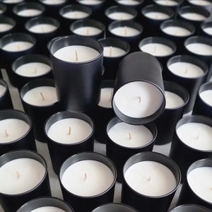 May include: A close-up view of a large quantity of black candle jars with white wax and wicks. The candles are arranged in a grid pattern.