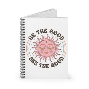 Puede incluir: Cuaderno de espiral blanco con una ilustración de sol retro en tonos rosa y marrón. El texto "BE THE GOOD SEE THE GOOD" está impreso alrededor del diseño del sol, con un arco iris.