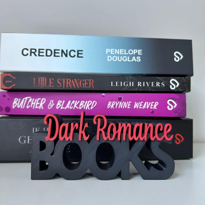 Op de afbeelding: Een stapel van vier boeken met titels als "Credence" en "Little Stranger". Een zwart en rood "Dark Romance BOOKS" bord staat voor de boeken. De boeken hebben verschillende kleuren, waaronder zwart, paars en blauw.