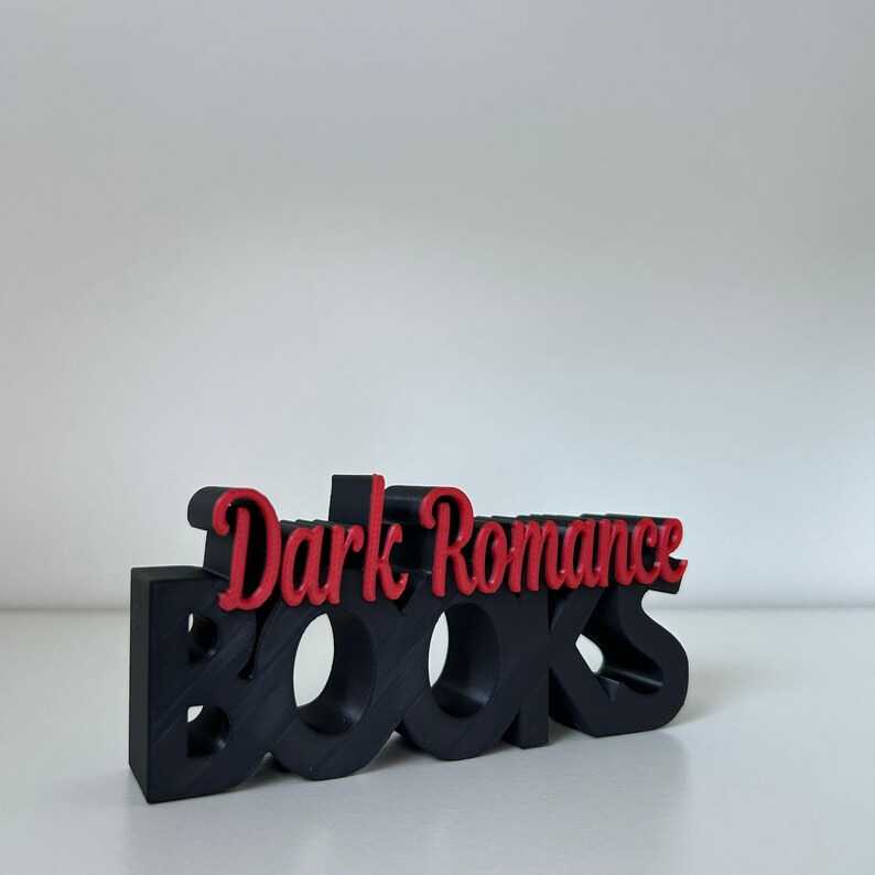 May include: A black 3D-printed sign with the words "Dark Romance Books" in a stylized font. The word "Books" is in black, and "Dark Romance" is in red. The sign is on a white surface.